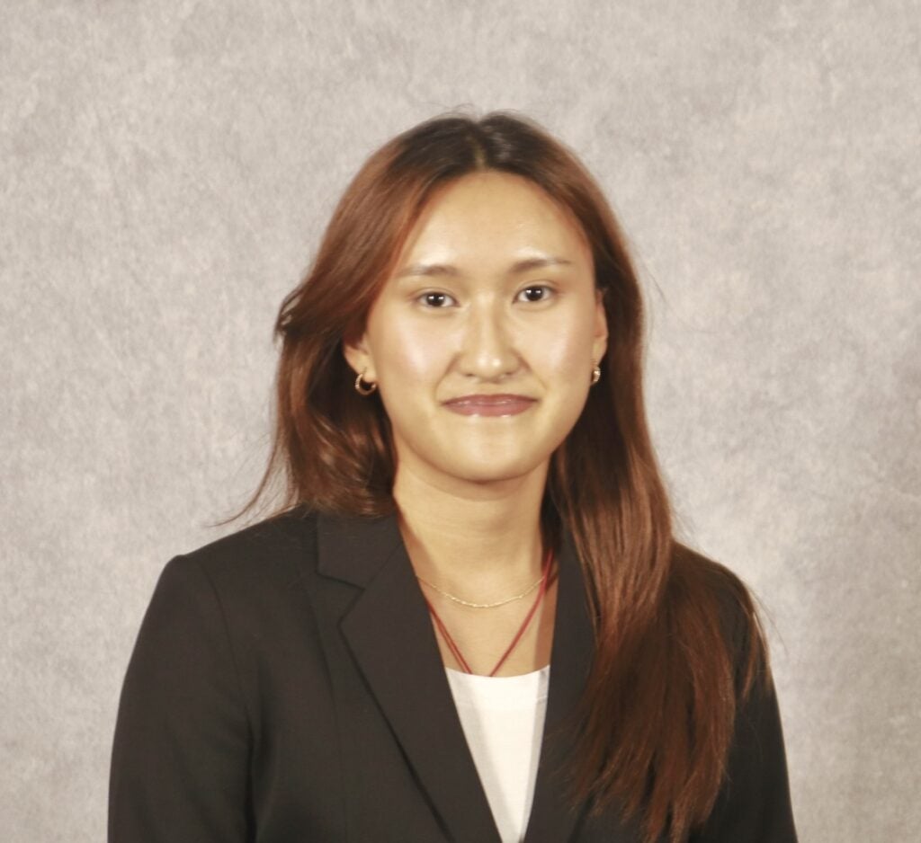 A woman with long brown hair wearing a black blazer and white top stands in front of a light gray backdrop, looking at the camera with a slight smile.