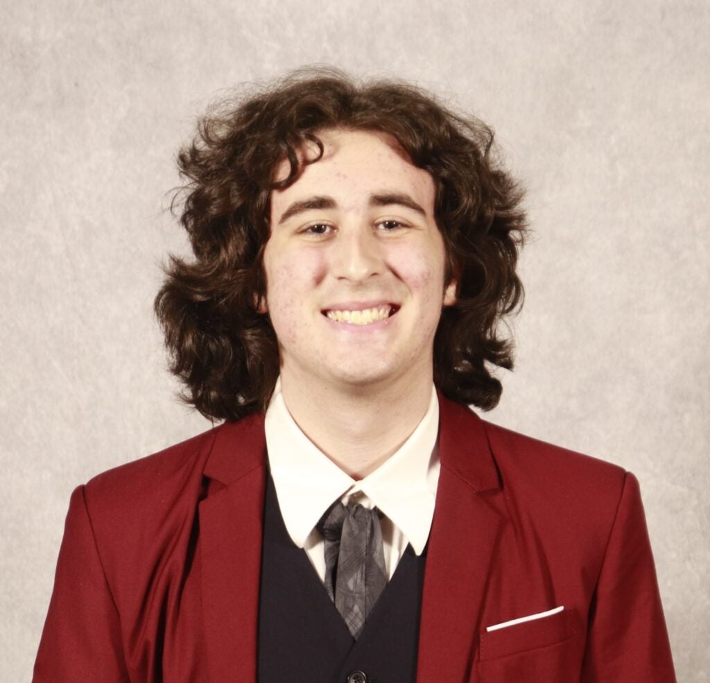 A young person with medium-length curly hair, wearing a red suit jacket, white shirt, and tie, smiles at the camera against a plain light gray background.