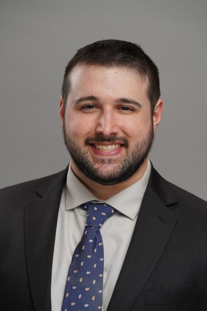 A man with short dark hair and a beard, wearing a dark suit, light shirt, and patterned tie, smiles in front of a plain gray background.