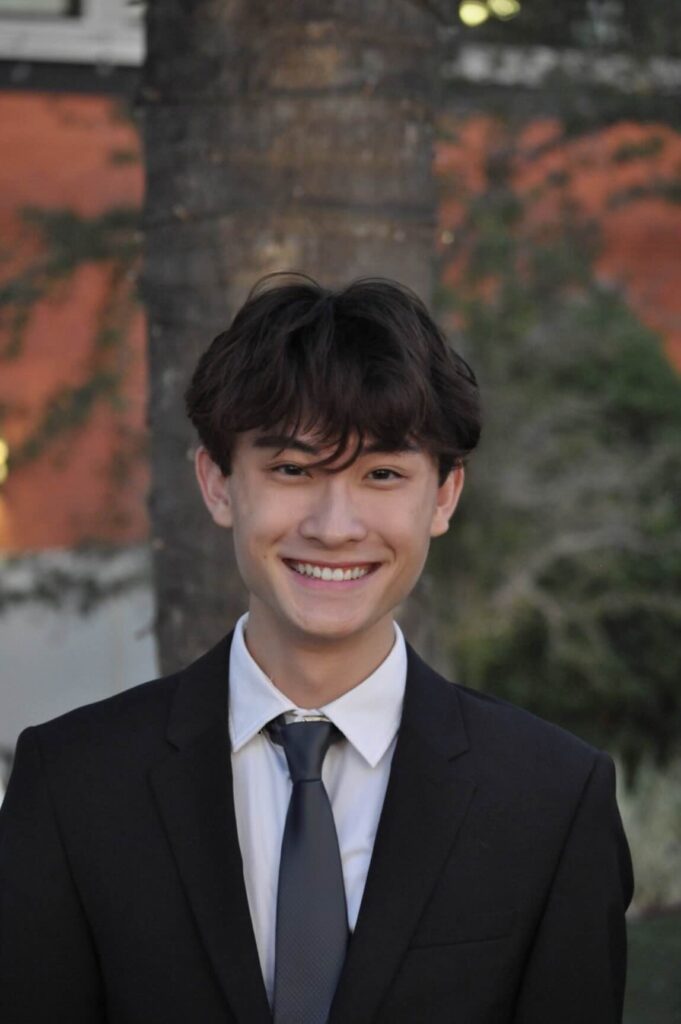 A young person with short, dark hair wearing a black suit, white shirt, and dark tie smiles in front of a tree and building.