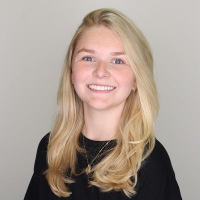 A young woman with long blonde hair, wearing a black top, smiles at the camera against a plain light-colored background.