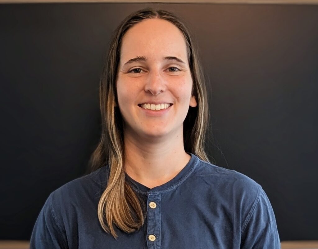 A person with long brown hair, wearing a blue shirt, is standing in front of a dark background and smiling at the camera.