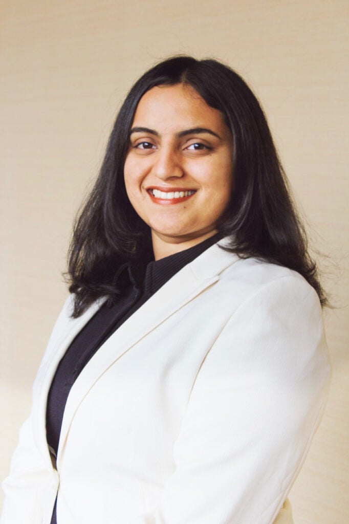 A woman with shoulder-length dark hair wearing a white blazer over a black top, smiling, posed against a neutral background.