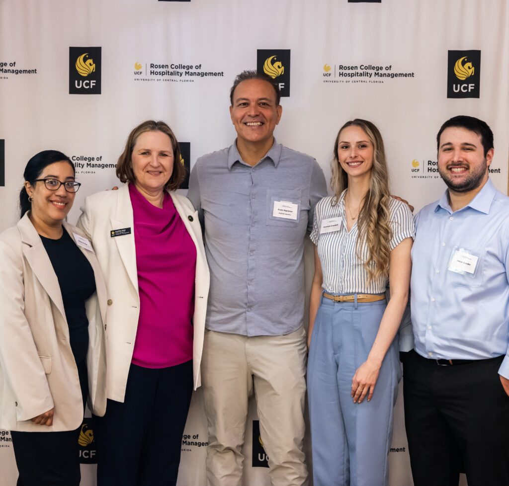 Five people stand and smile in front of a Rosen College of Hospitality Management and UCF step-and-repeat banner, wearing business or business casual attire.