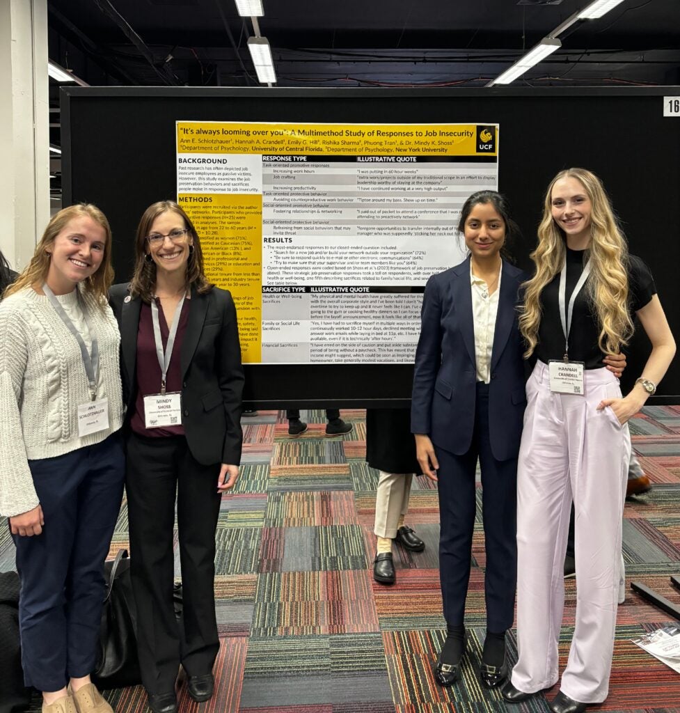 Four women stand in front of a research poster on job insecurity at a conference, smiling at the camera.