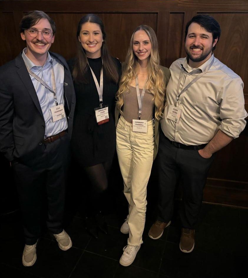 Four people wearing conference name tags stand together, smiling, in business casual attire against a dark wooden wall.