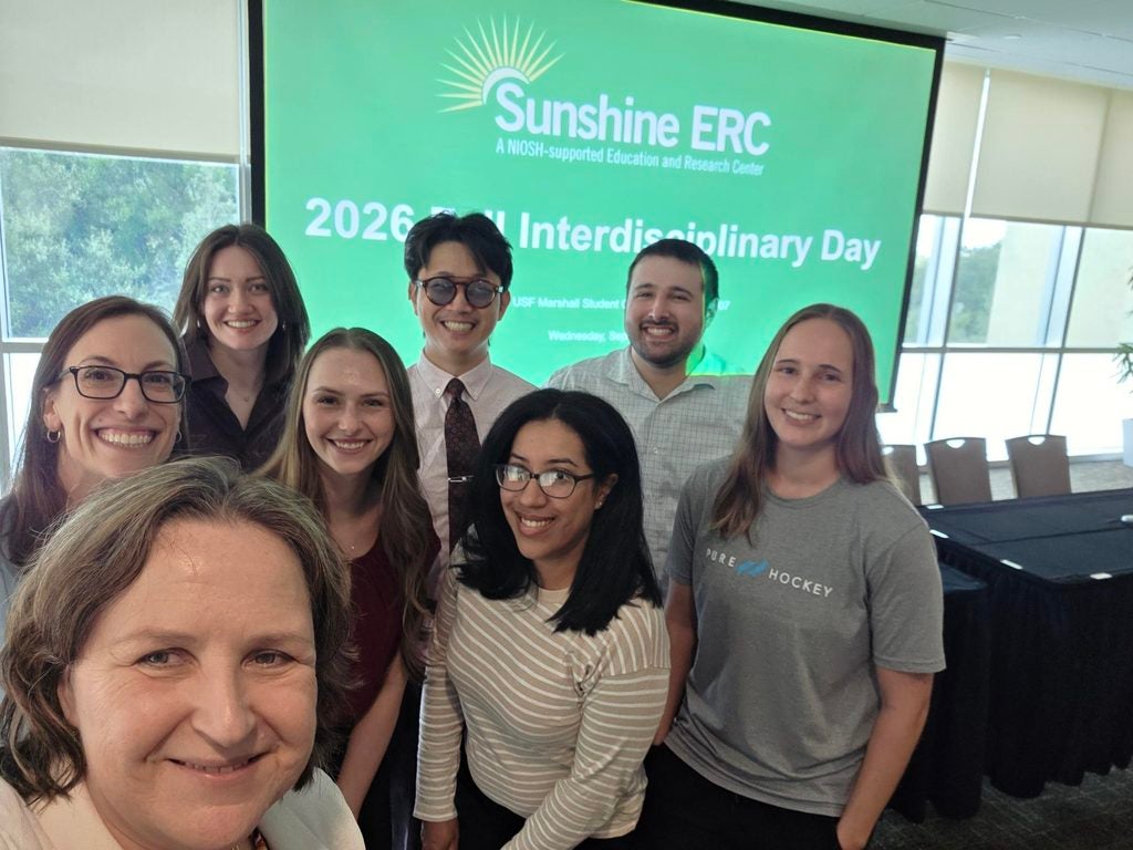Eight people stand indoors smiling for a group selfie in front of a screen displaying “Sunshine ERC 2026 Interdisciplinary Day” and other text.