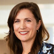 A woman with brown hair and hoop earrings smiles at the camera, wearing a blue patterned top, with a blurred indoor background.