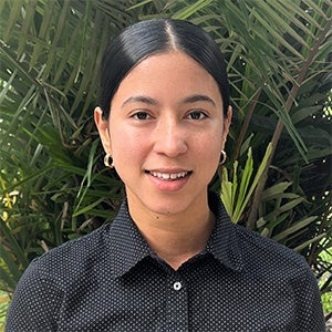 A person with long dark hair, wearing a black patterned shirt and gold hoop earrings, stands in front of green foliage.