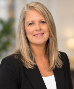 A woman with long blonde hair, wearing a black blazer and white top, smiles at the camera against a softly blurred indoor background.