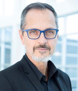 A man with short gray hair, a goatee, and blue eyeglasses wearing a black suit jacket stands in a bright, modern indoor setting.