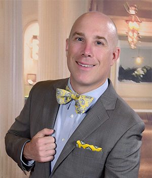 Bald man in a gray suit with a yellow patterned bow tie and pocket square, standing indoors with a slight smile.