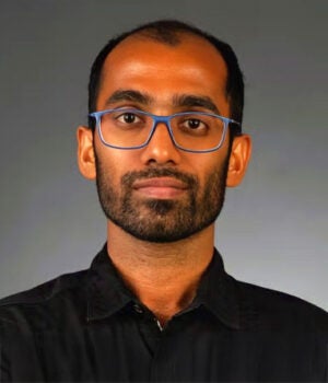 A man with a beard and blue glasses wearing a black collared shirt is facing the camera against a plain gray background.