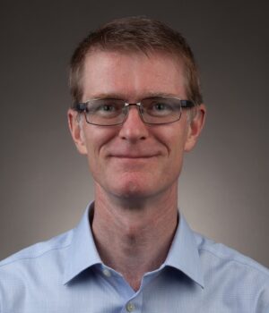 A man with short brown hair and glasses, wearing a light blue collared shirt, is facing the camera against a plain background.