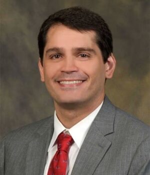 A man in a gray suit, white shirt, and red tie is smiling at the camera against a neutral, blurred background.