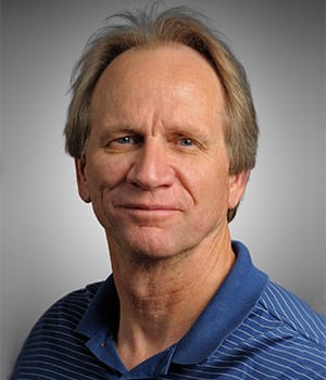 Middle-aged man with light hair wearing a blue striped polo shirt, posing in front of a neutral gray background.