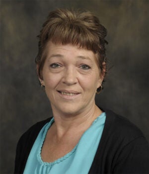 A middle-aged woman with short brown hair, wearing a light blue top and black cardigan, poses in front of a plain dark background.