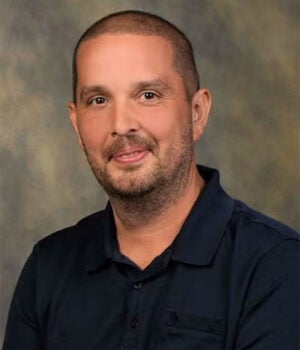 A man with short hair and a trimmed beard wearing a dark collared shirt sits in front of a mottled, neutral-colored background.