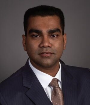A man in a dark suit, white shirt, and patterned tie poses for a professional headshot against a plain gray background.