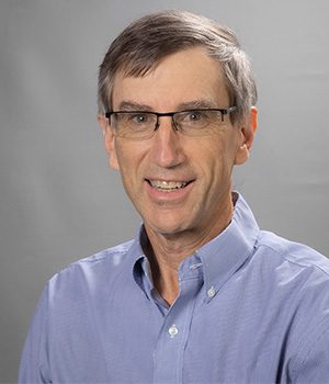 A middle-aged man with short gray hair and glasses, wearing a light blue button-up shirt, smiles at the camera against a plain gray background.