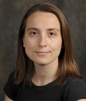 A woman with straight, shoulder-length brown hair and a black top poses in front of a dark, blurred background.