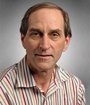 A middle-aged man with short brown hair wearing a striped collared shirt, posed in front of a plain gray background.