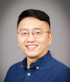 A person with short black hair, glasses, and a blue button-up shirt smiles in front of a plain gray background.