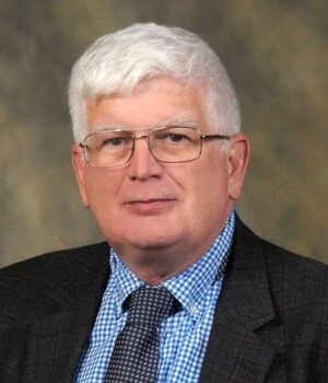 Older man with short white hair and glasses, wearing a dark jacket, blue checkered shirt, and a patterned tie, posed in front of a neutral background.