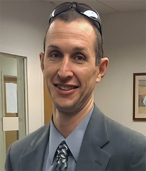 A man in a gray suit and patterned tie smiles at the camera while wearing sunglasses on his head in an office setting.