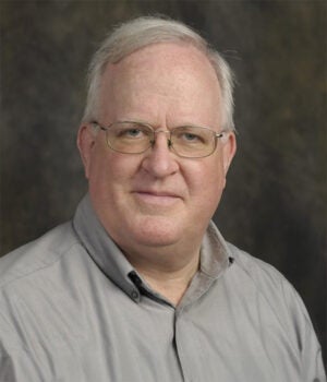 A middle-aged man with gray hair and glasses, wearing a light gray collared shirt, poses against a dark, neutral background.