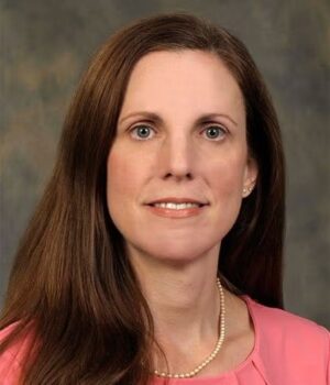A woman with long brown hair, wearing a pink blouse and a pearl necklace, poses against a neutral background.