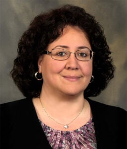 A woman with curly dark hair, glasses, and hoop earrings is wearing a patterned blouse and dark blazer, posing in front of a neutral background.