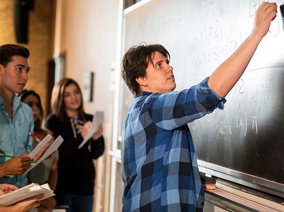 A student writes on a chalkboard while others stand nearby, observing and taking notes in a classroom setting.