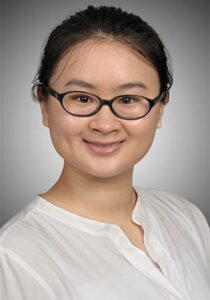 A person with glasses and dark hair tied back, wearing a white blouse, smiles against a gray gradient background.