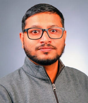 A man with short black hair, beard, and glasses wears a grey zip-up sweater and looks at the camera against a plain grey background.