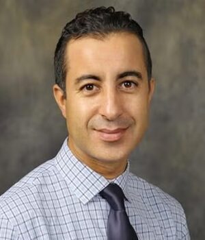 Man with short dark hair wearing a checkered shirt and dark tie, posing in front of a neutral, mottled background.