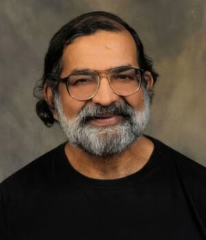 A middle-aged man with glasses, gray beard, and mustache is smiling slightly, wearing a black shirt against a neutral background.