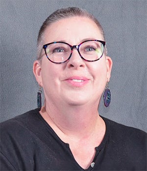 A person with short hair, glasses, and earrings, wearing a dark shirt, is smiling in front of a plain gray background.