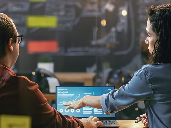 Two people review data on a laptop screen displaying a graph and the word "Analysis" in an office setting with charts on a glass wall behind them.
