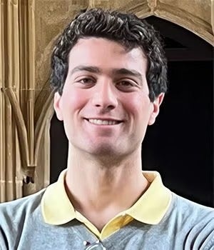 A young man with short, curly hair smiles, wearing a grey sweater over a yellow collared shirt, standing in front of a stone archway.