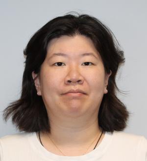 A person with shoulder-length dark hair and a neutral expression is facing the camera against a plain light background.