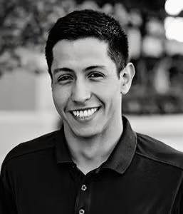 A man with short dark hair, wearing a dark polo shirt, smiles while looking at the camera in an outdoor setting.