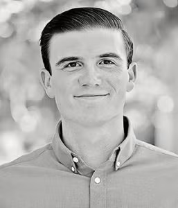 A young man with short, neatly styled hair and a button-up shirt smiles slightly while looking at the camera. The background is blurred.