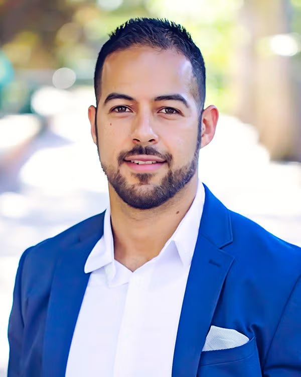 A man with short dark hair and a trimmed beard wears a blue suit jacket, white shirt, and pocket square, standing outdoors with a blurred background.