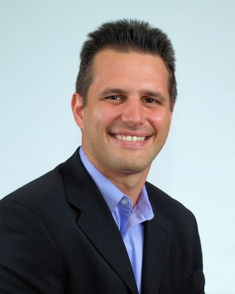 A man in a dark suit jacket and light blue shirt smiles at the camera against a plain light background.