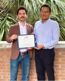 Two men stand side by side outdoors; one holds a certificate from ASA framed in blue. Both are facing the camera with a brick wall and green foliage in the background.