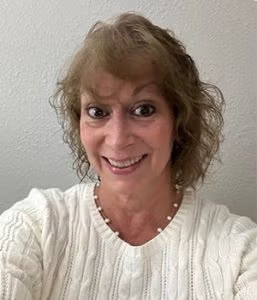 A woman with curly light brown hair wearing a white sweater and a beaded necklace smiles at the camera against a light-colored wall.