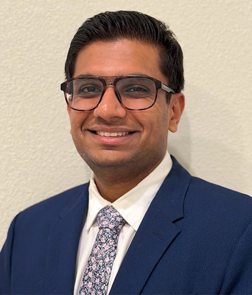 A man wearing glasses, a navy blue suit, white shirt, and floral tie, smiling in front of a plain light-colored wall.
