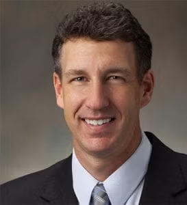 A man with short brown hair wearing a dark suit, white shirt, and striped tie, smiling at the camera against a neutral background.
