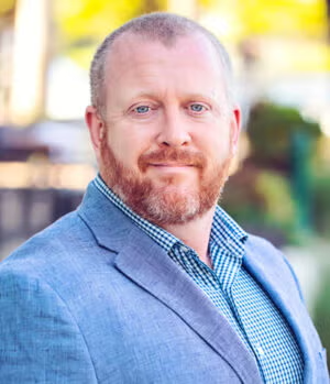 A man with a short beard and short hair, wearing a light gray blazer over a checkered shirt, poses outdoors with a neutral expression.